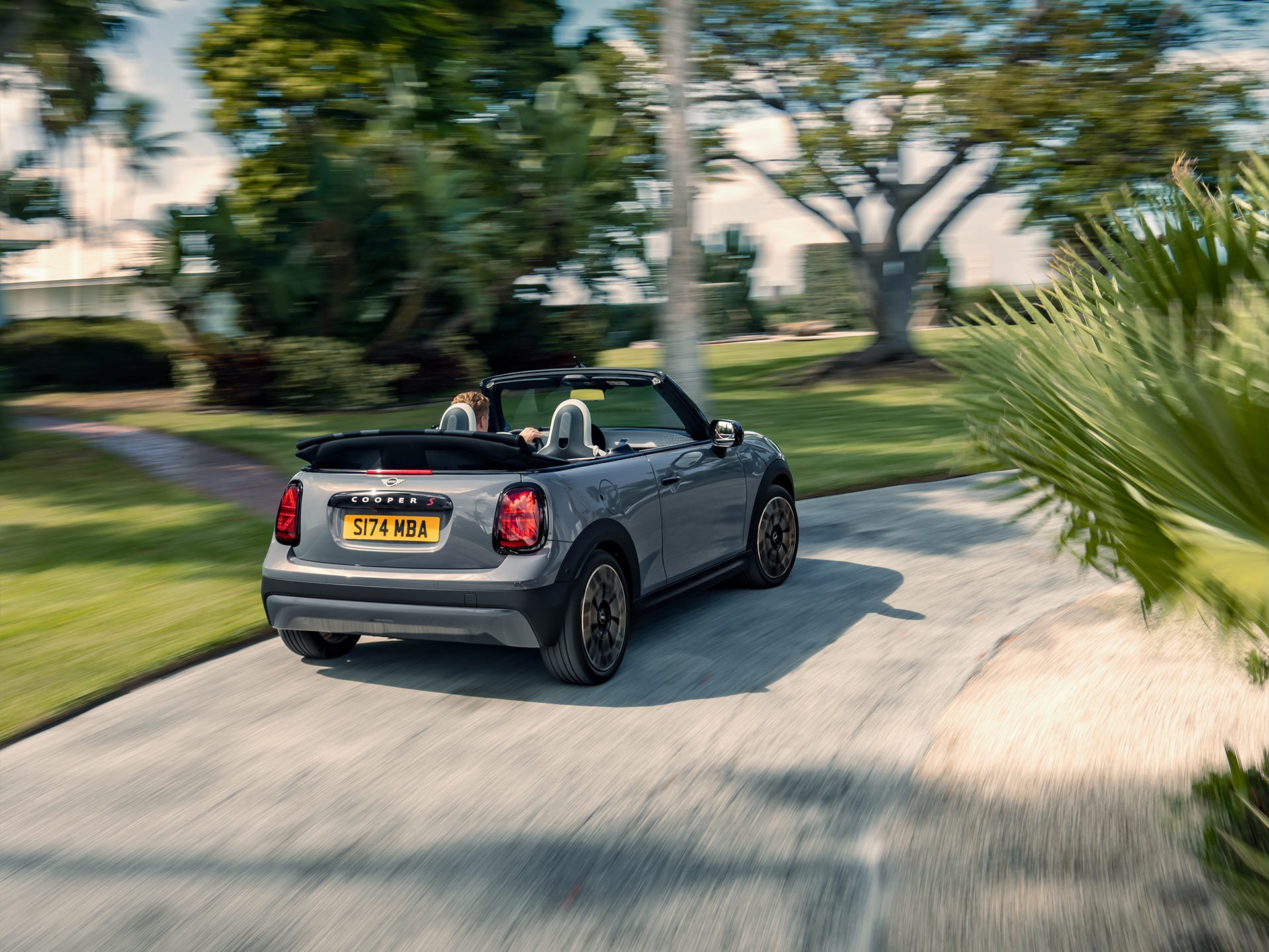 Angled back view of the new MINI Cooper Convertible in motion with the top down and a blurred, lush backdrop.